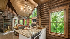 A log cabin kitchen with a large island, granite countertop, and a view of the forest through a large window. - Romance-Grande Old World-Lux Hot Tub-Kayaks-Private Spa w/ Massage Chair & Sauna (Logan)