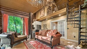 A spacious living room with a large sofa, a smaller armchair, and a red patterned rug. A staircase with ornate railings is visible in the background. - Romance-Grande Old World-Lux Hot Tub-Kayaks-Private Spa w/ Massage Chair & Sauna (Logan)
