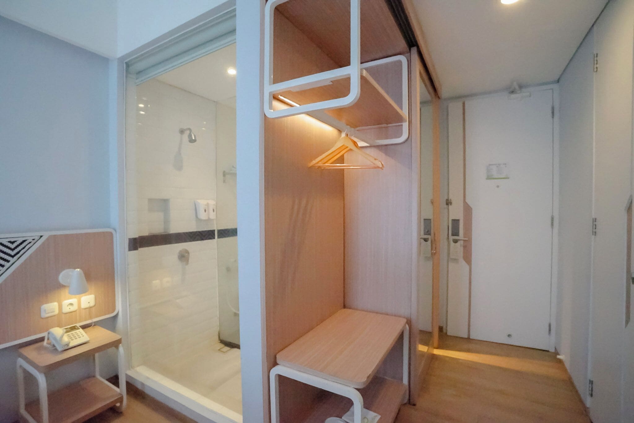 A modern hotel room with a glass shower enclosure, a wooden bench, a small table with a lamp, and a coat rack.