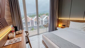 A hotel room with a bed, a desk with a chair, a table with a lamp, and a view of buildings through large windows. - Facade Hotel by Azana (Tawangmangu)