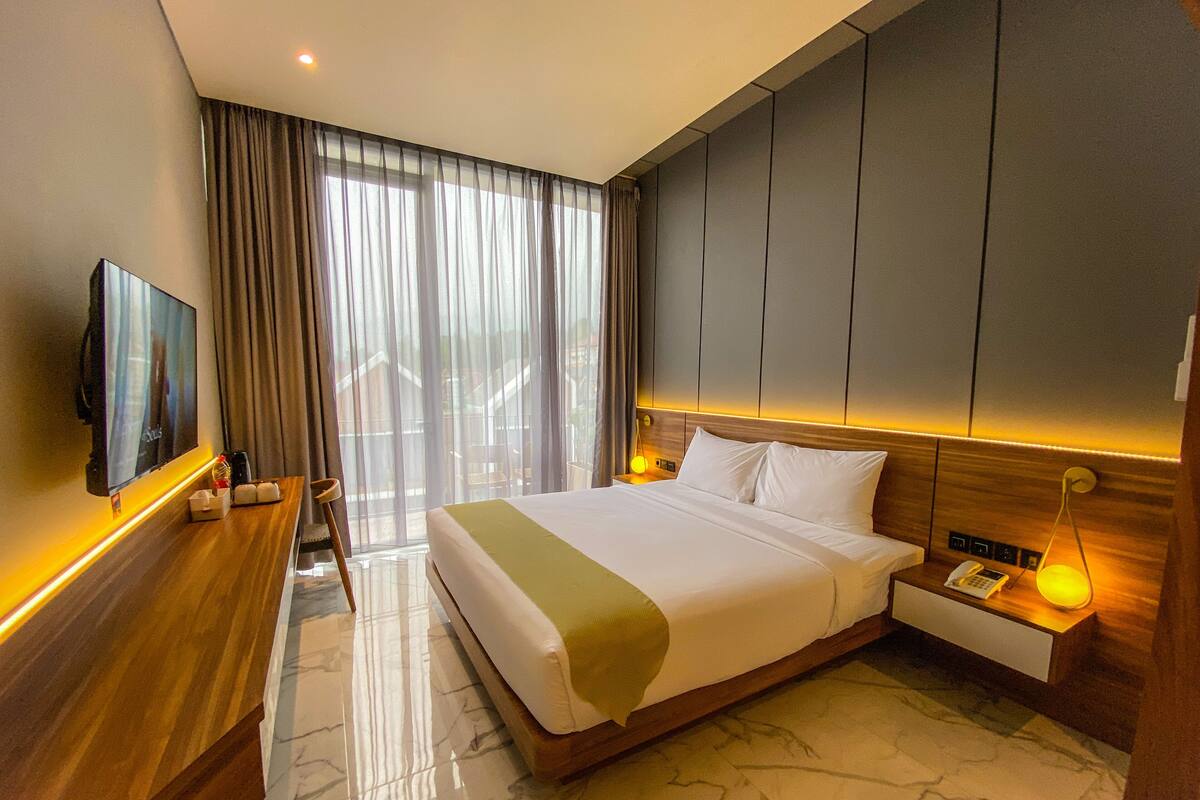 A hotel room with a large bed, a TV mounted on the wall, a bedside table with a lamp, and a view of the outdoors through the window.