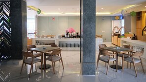 A modern dining area with marble flooring, wooden tables and chairs, and a central island with kitchen appliances and a pink flower arrangement. - Facade Hotel by Azana (Tawangmangu)