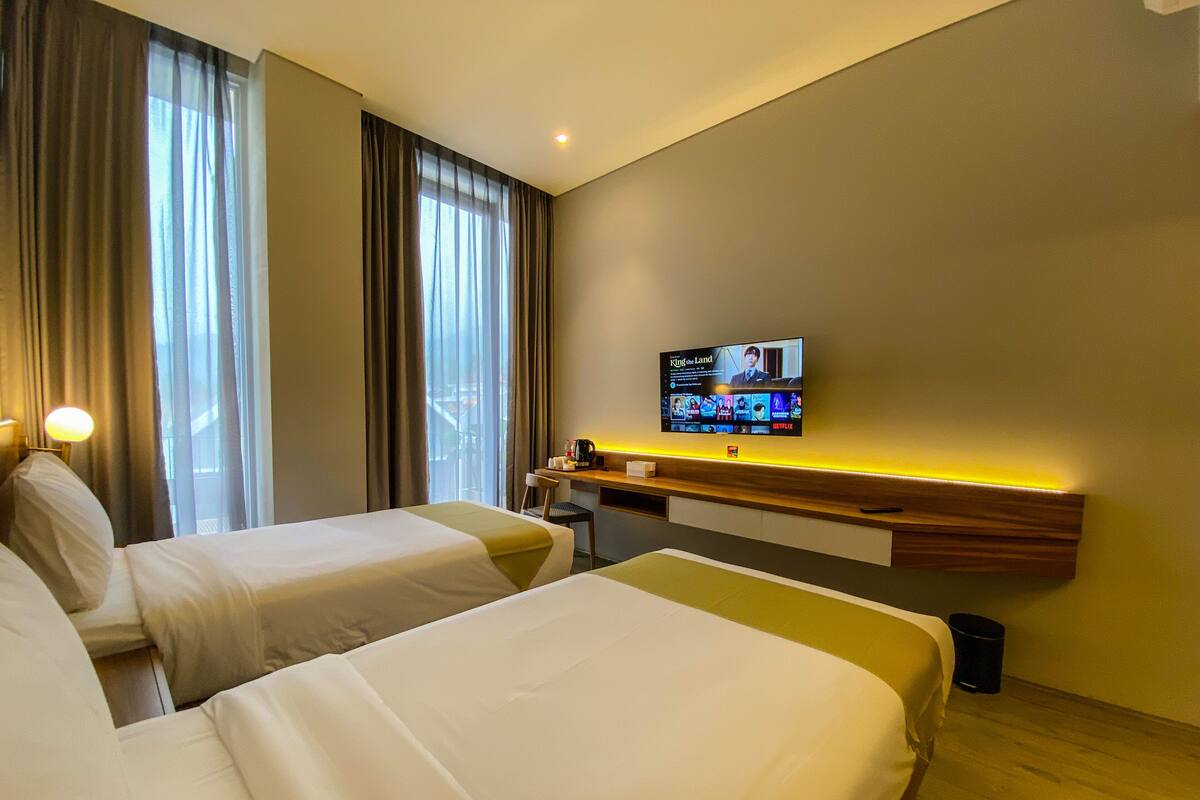 A hotel room with two beds, a desk, a television displaying a movie, and a window with curtains.