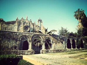 An ancient, weathered building with arched doorways and statues, surrounded by greenery and palm trees. - Interno 10 (Syracuse)