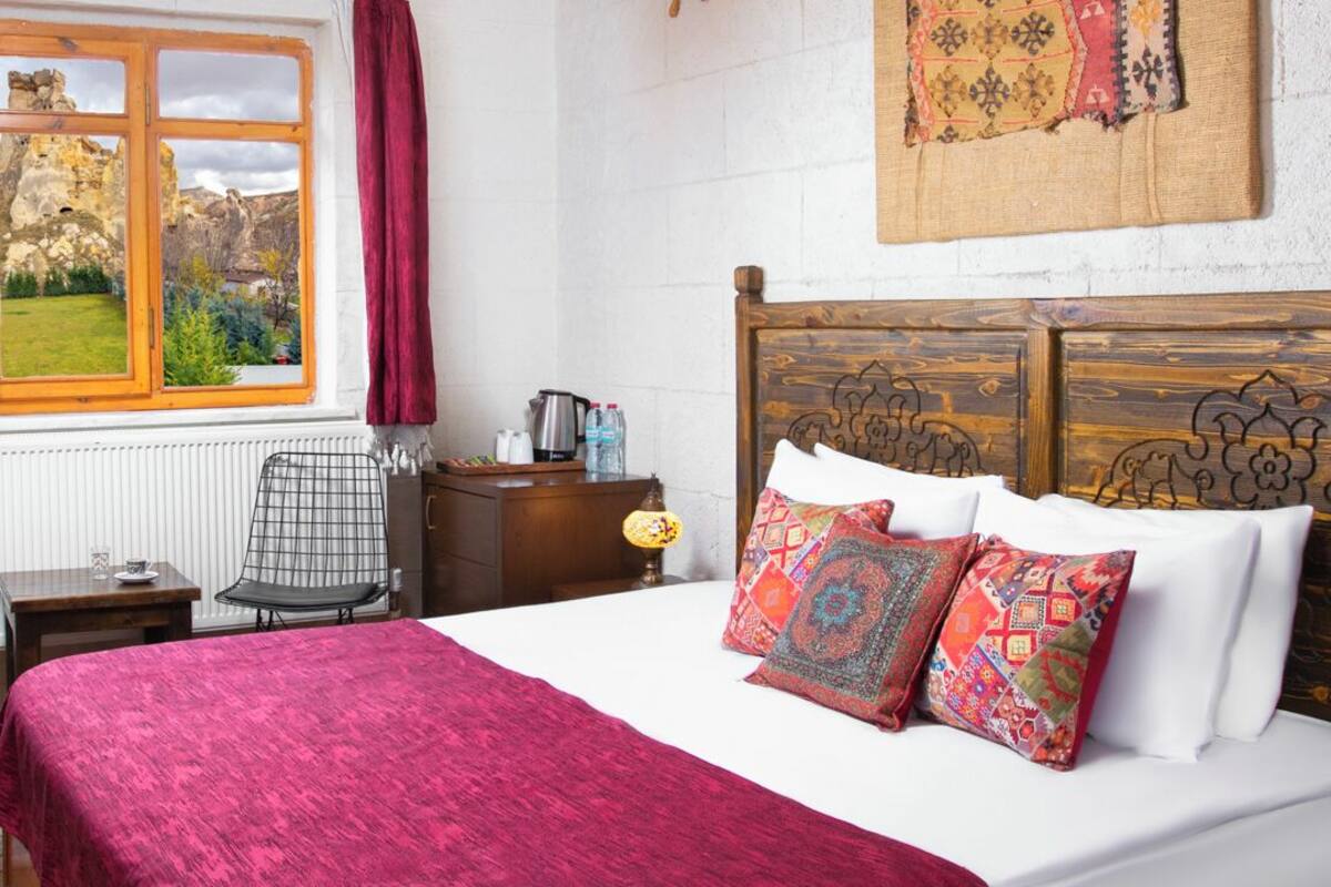 A bedroom with a bed, a wooden headboard, a window with a view of a mountainous landscape, a chair, a small table, and a lamp.
