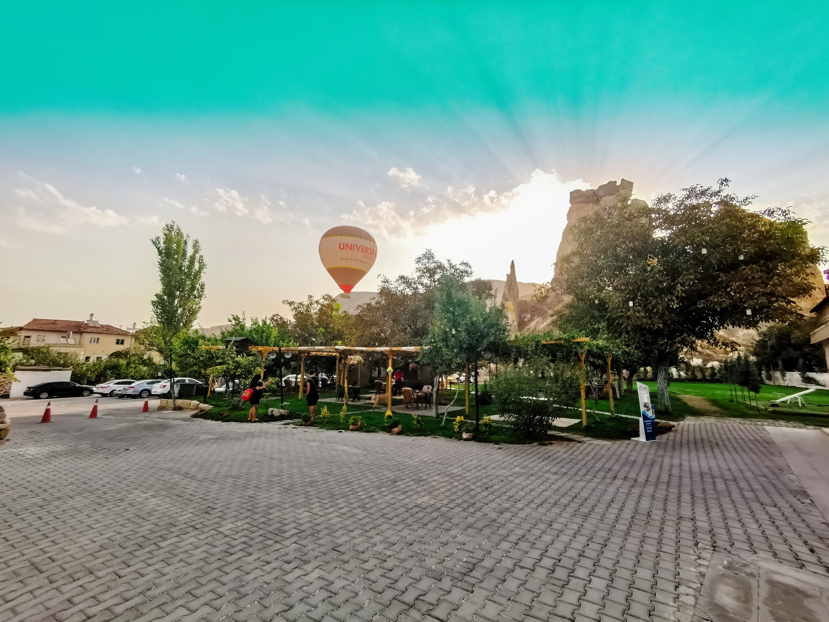 Un patio empedrado con un globo aerostático al fondo, rodeado de árboles y edificios.