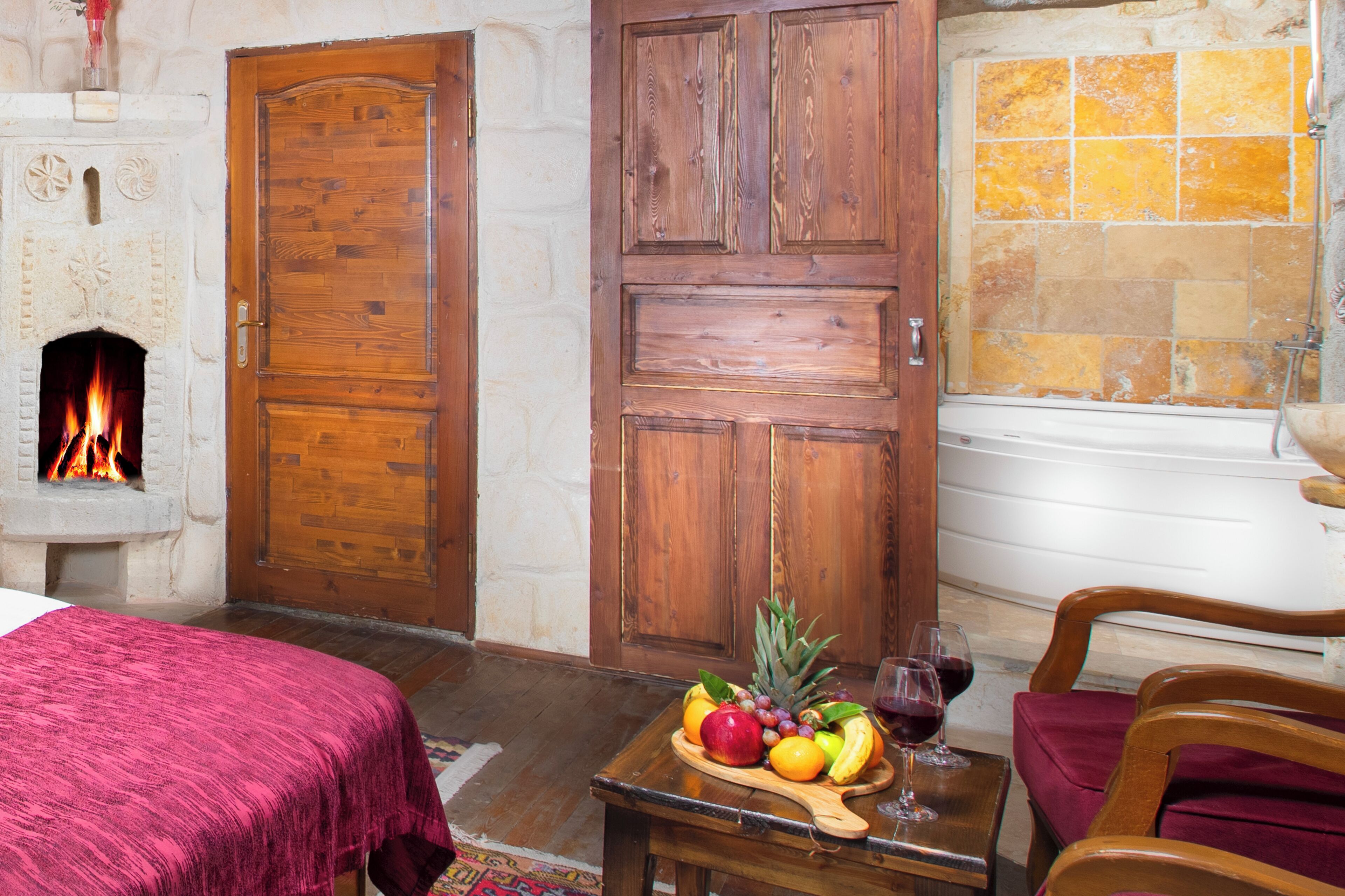 A hotel room with a fireplace, a wooden door, a fruit and wine display, and a red upholstered chair.