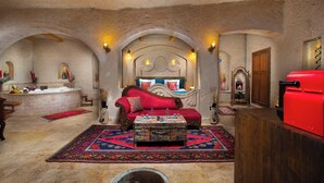 A room with a stone ceiling, a red sofa, a patterned ottoman, a red grill, and a stone fireplace.