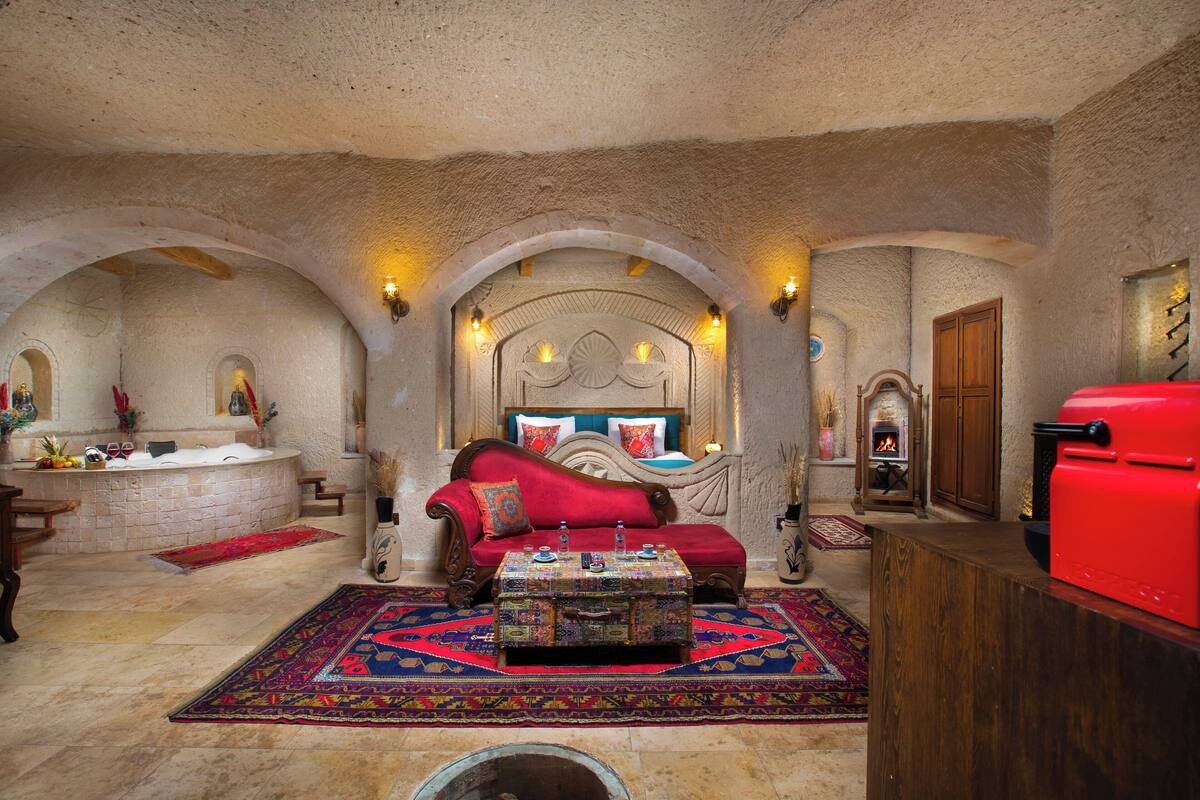 A room with a stone ceiling, a red sofa, a patterned ottoman, a red grill, and a stone fireplace.
