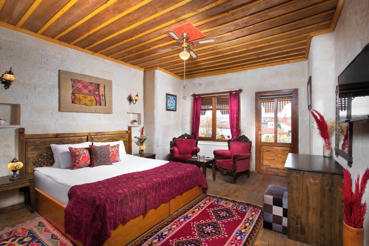 A bedroom with a wooden ceiling, a bed with a red blanket, a small table, two chairs, and a view of the outside through a window.