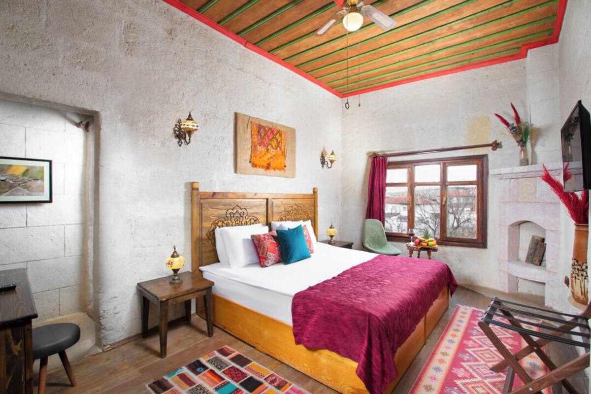 A bedroom with a wooden bed, a nightstand, a chair, a window with curtains, a fireplace, and a wall-mounted TV.