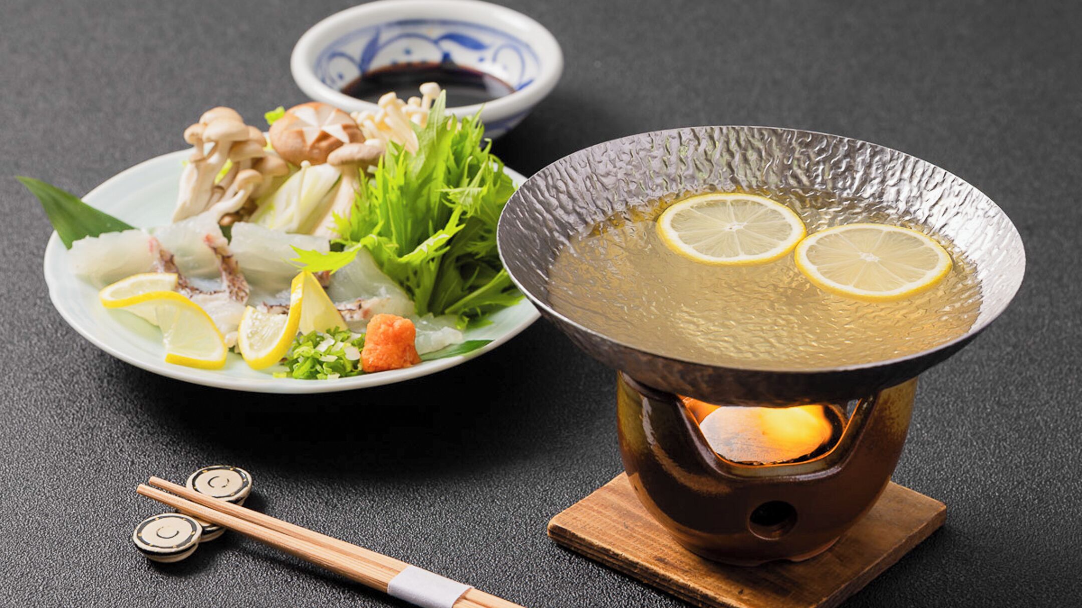 Una olla humeante con rodajas de limón, un plato de sashimi y palillos sobre una mesa.