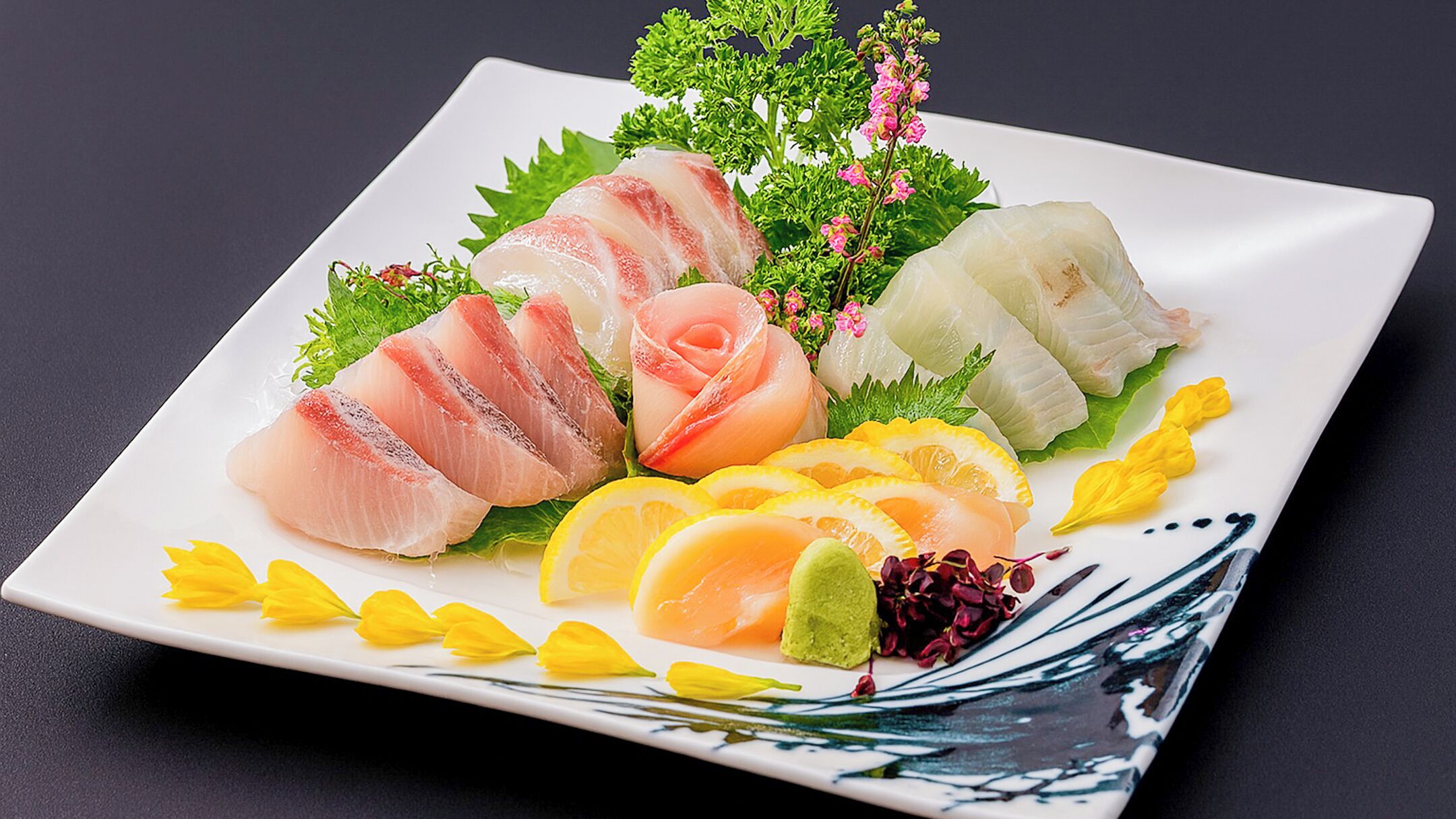 A square plate with various sashimi pieces, garnishes, and decorative elements.