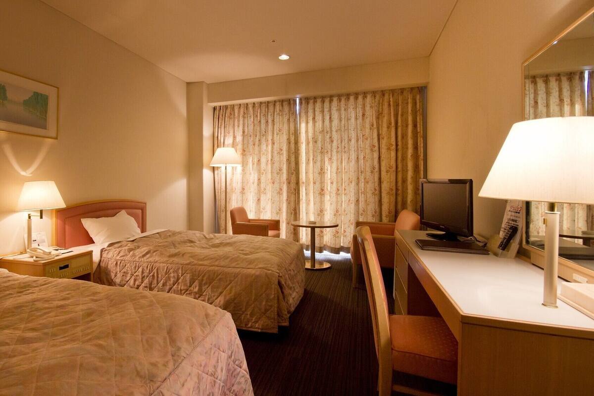 A hotel room with two beds, a desk, a chair, a television, and a lamp.