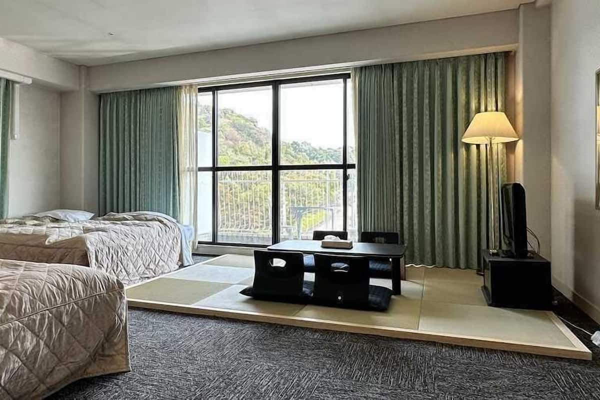 A hotel room with two beds, a small table, a TV, and a view of a mountainous landscape through large windows.