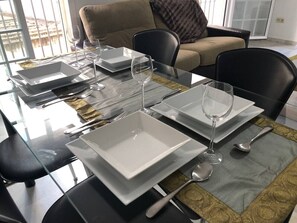 A dining table set with white plates, glasses, and cutlery, with a view of a balcony in the background.