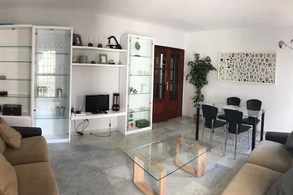 A modern living room with a glass coffee table, a TV, and a dining area with chairs and a table.