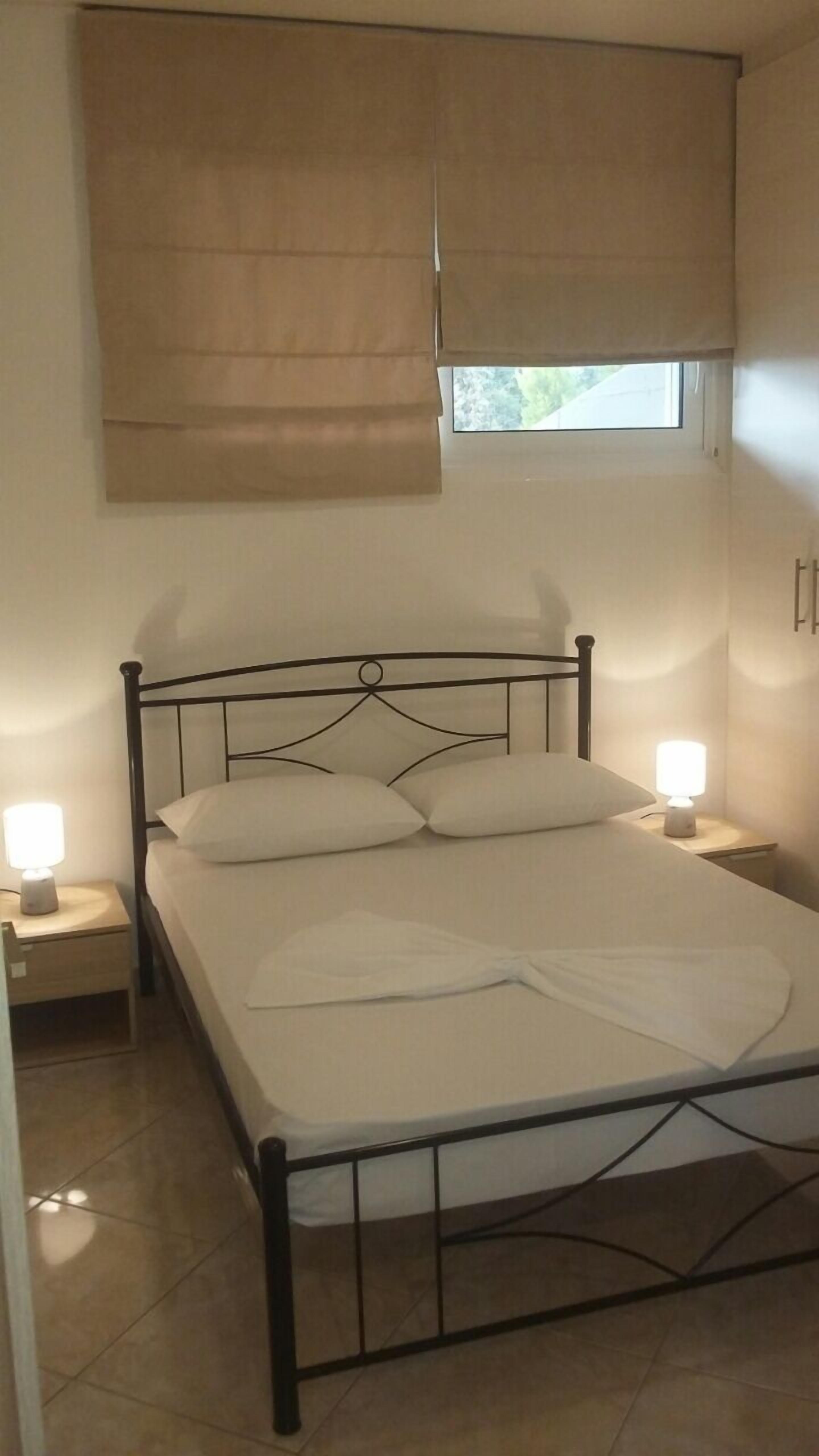 Un dormitorio con estructura de cama metálica, dos mesitas de noche con lámparas, una ventana con persiana y una pequeña mesita auxiliar.