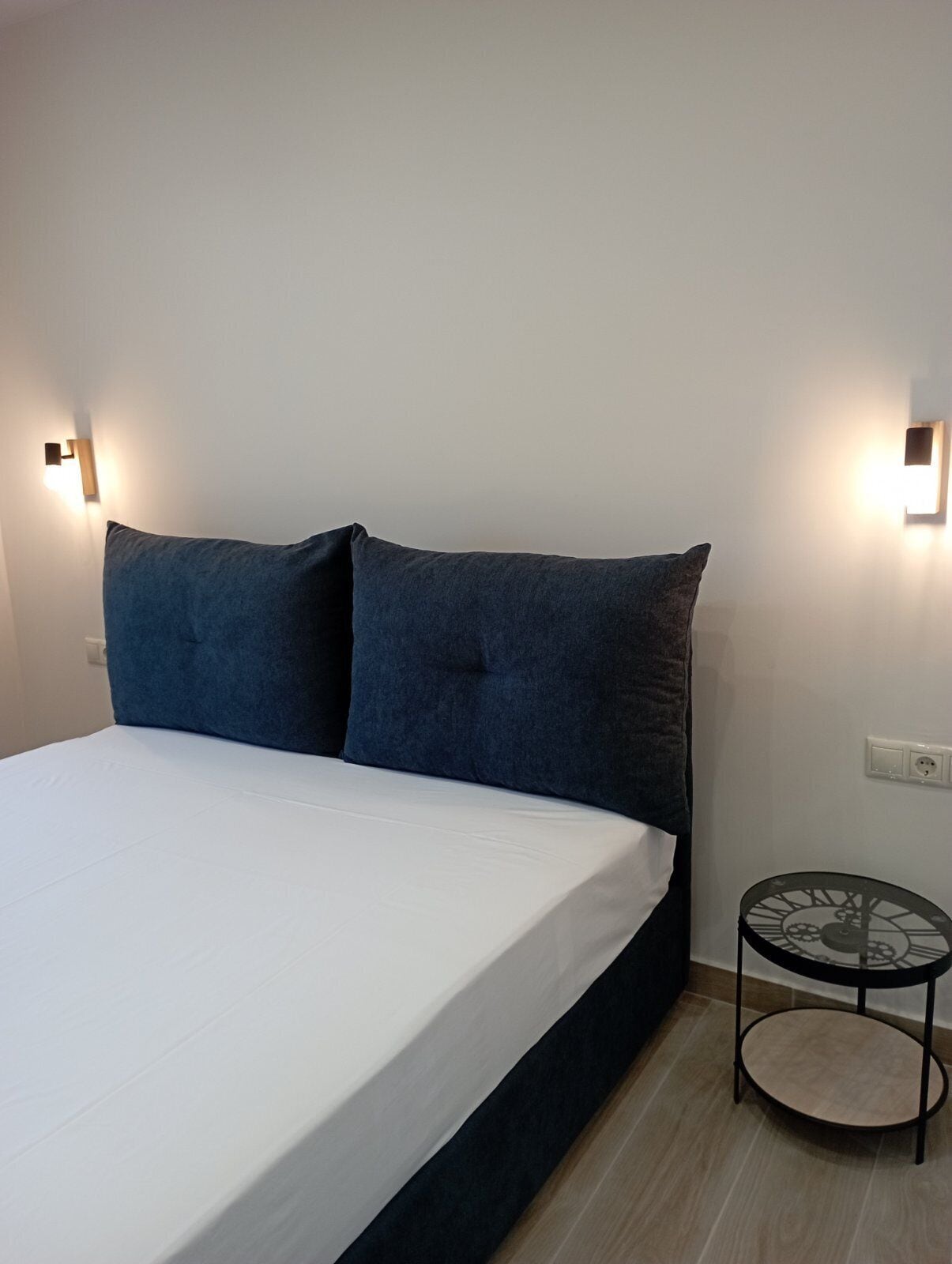 A bed with a white mattress and two dark blue pillows, a small round table with a black metal frame, and two wall-mounted light fixtures.