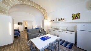 Une cuisine de taille réduite avec des meubles blancs, un évier et un réfrigérateur. On y trouve une table à manger avec des chaises et un set de table bleu. Un canapé et un téléviseur sont visibles au fond.
