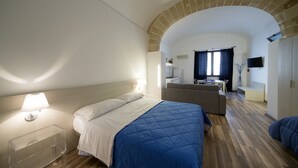 Une chambre d’hôtel avec un lit, une table de chevet dégagée, un canapé, une télévision et, au fond, une kitchenette.