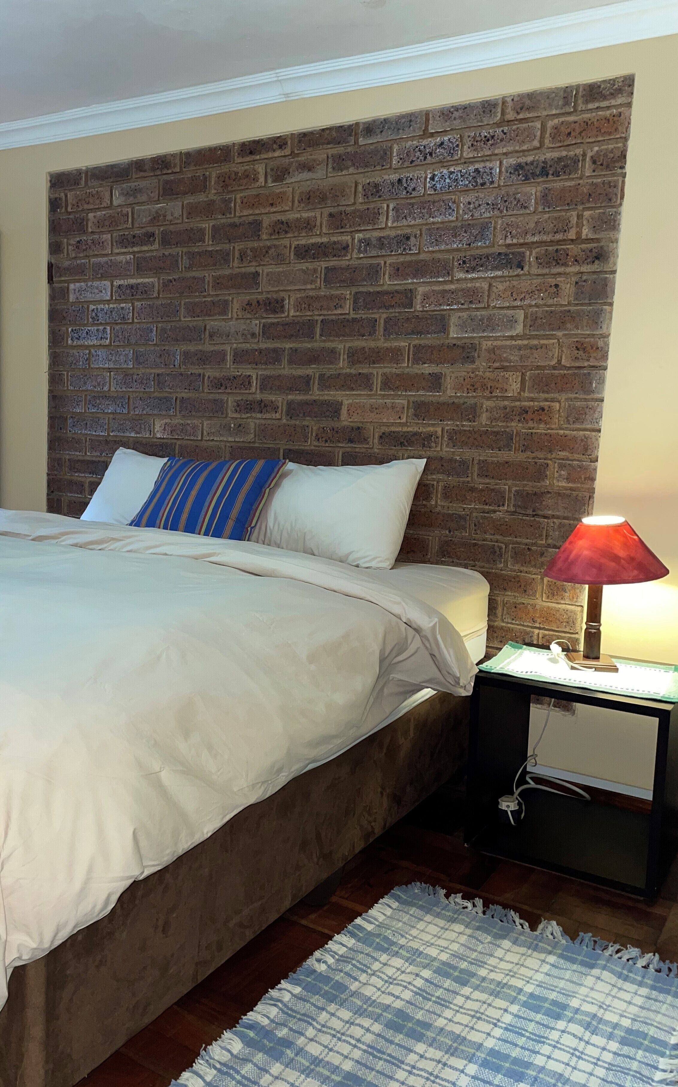 A bedroom with a brick headboard, a bed with white bedding, a striped pillow, a red lamp on a bedside table, and a blue and white checkered rug.