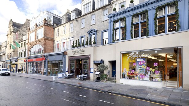 A street view with a row of shops, including a cafe and a store with a bright display window. - JOIVY 5-star Luxurious George Street Apartment (Edinburgh)
