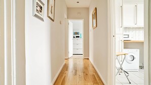 A hallway with wooden flooring, white walls, and framed pictures. There is a door leading to a kitchen area with white cabinets and a washing machine. - JOIVY Bright Royal Mile Apartment (Edinburgh)