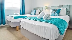 A hotel room with two beds, each with a teal blanket and white bedding. There is a window with blue curtains, a wall-mounted lamp, and a floral artwork above the beds. - JOIVY Bright Royal Mile Apartment (Edinburgh)
