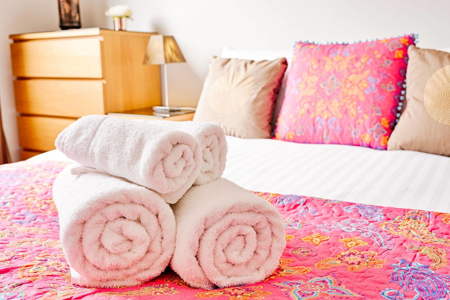 A neatly made bed with pink and white towels, a colorful patterned bedspread, and a wooden nightstand with a lamp.