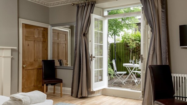 A room with a bed, two chairs, a table with a bottle and glasses, and a view of a garden through an open door. - JOIVY New Town Boutique Apartment on India Street (Edinburgh)
