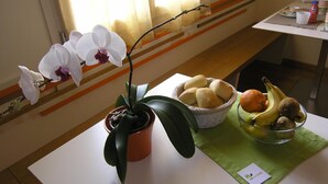 Une orchidée en pot, un panier de petits pains et un saladier de fruits sur une table.