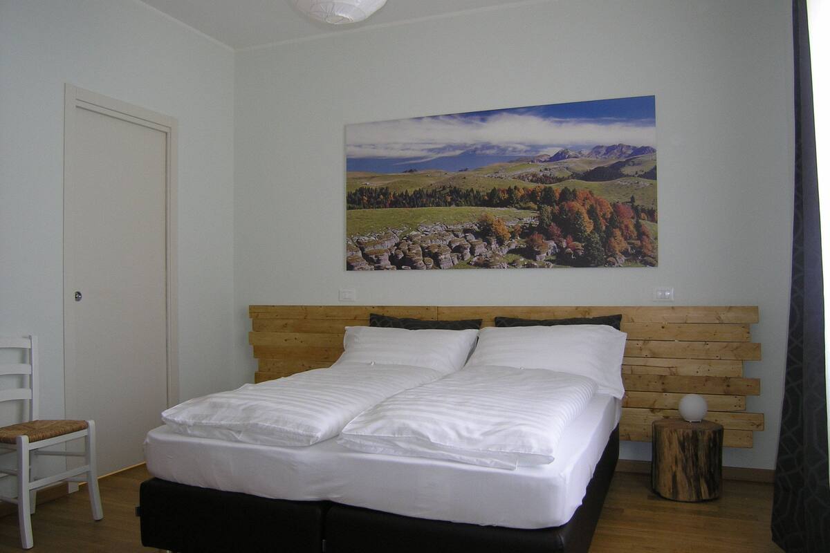 Une chambre à coucher avec une tête de lit en bois, un lit recouvert de linge de lit blanc, une chaise et un tableau représentant un paysage accroché au mur.