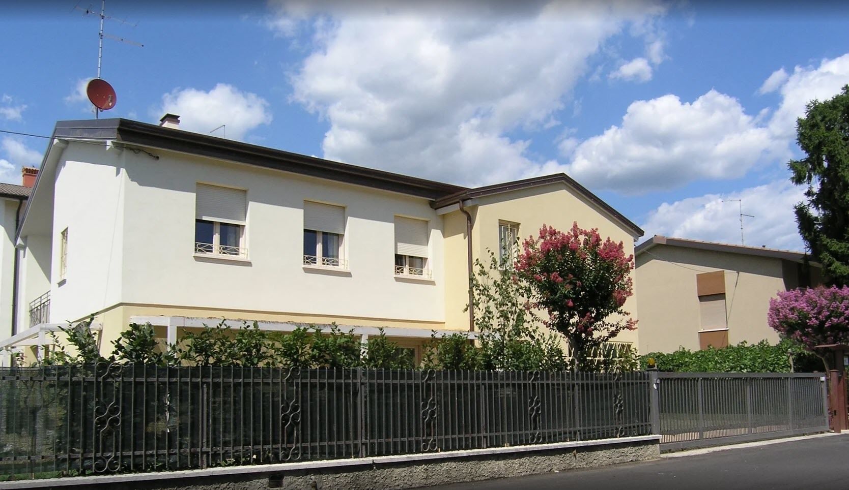 A two-story residential building with a beige facade, a black metal fence, and a tree with pink flowers in front.
