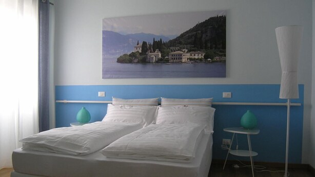 Un lit double avec du linge de lit blanc, des murs peints dans des tons de bleu et de blanc, un tableau représentant un paysage côtier et une petite table de chevet sur laquelle est posé un vase vert.