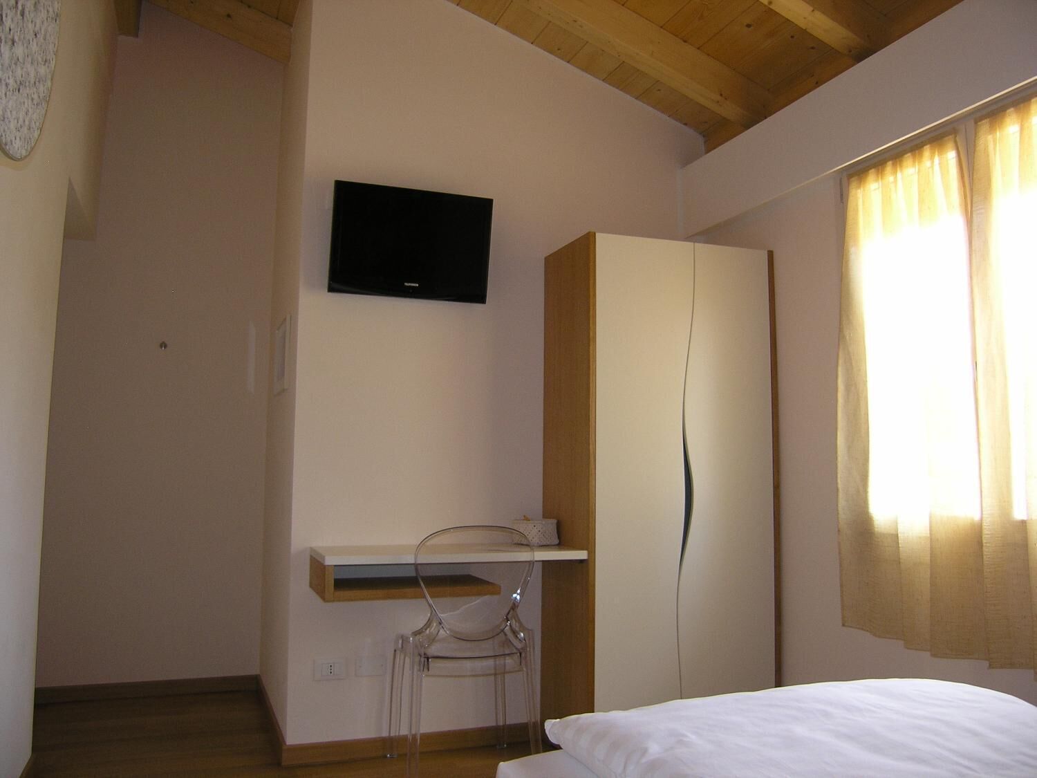 A bedroom with a bed, a TV mounted on the wall, a wooden wardrobe, and a desk with a chair.