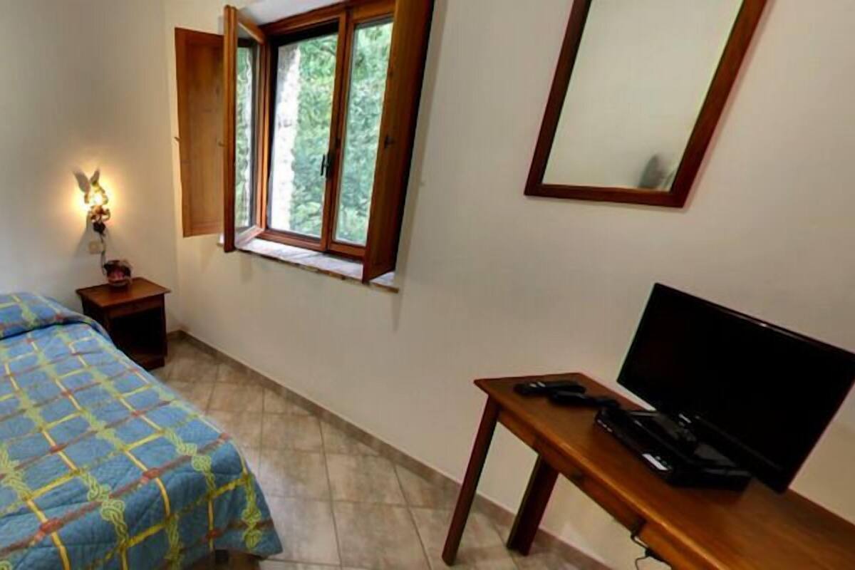 Un dormitorio con cama, una mesita de noche con lámpara, una ventana con persiana abierta y un mueble para televisión.