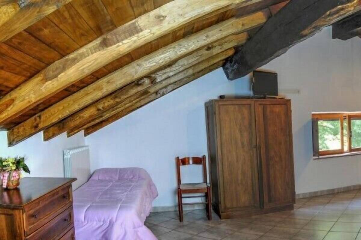 Un dormitorio con techo inclinado, vigas de madera, una cama, una silla, un armario y una ventana con vistas a la vegetación.