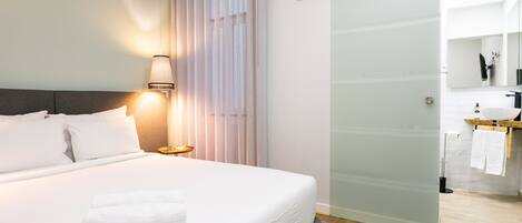 Una habitación de hotel moderna con una cama grande, una lámpara de noche, una puerta corredera hacia el baño y vistas al baño a través de la puerta.