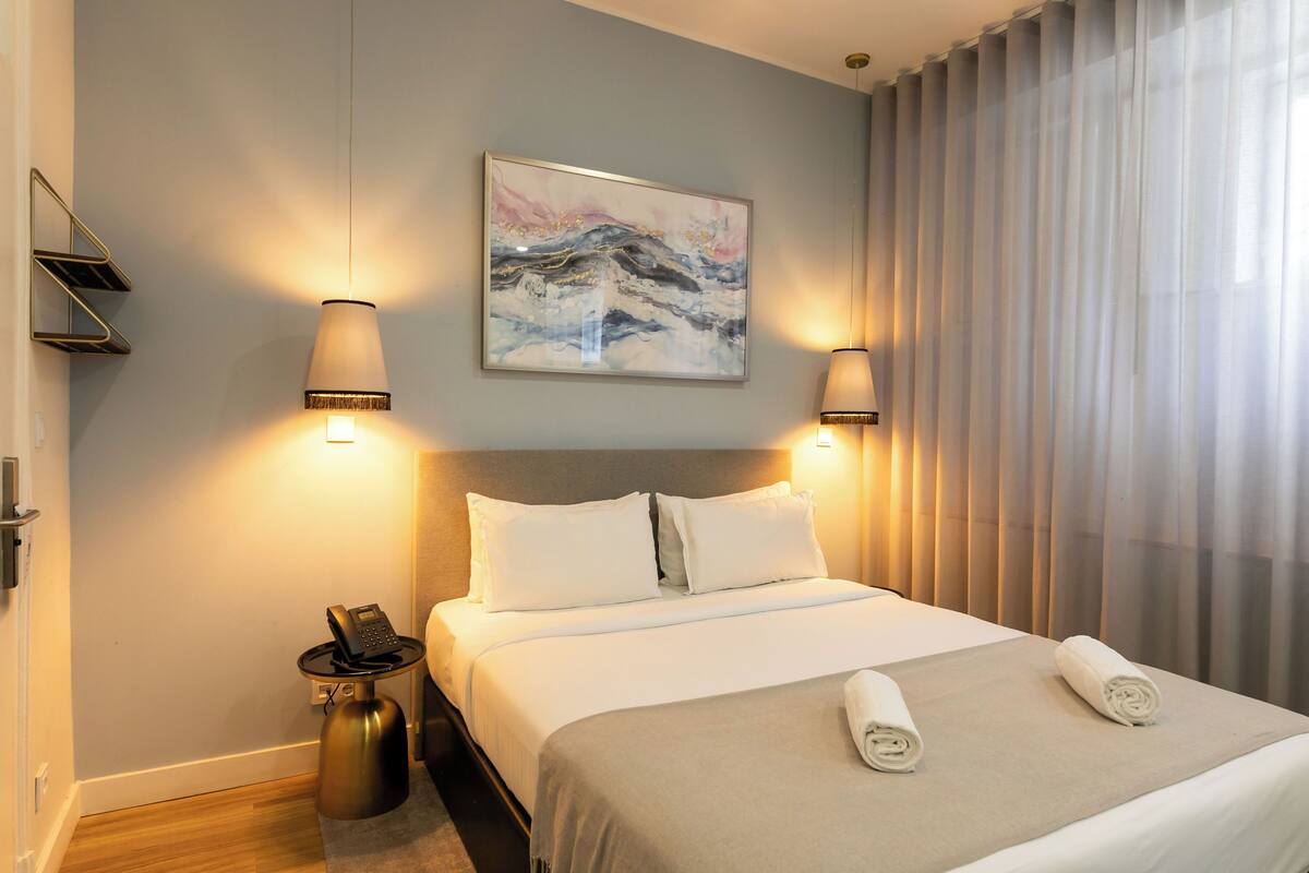A hotel room with a bed, two bedside lamps, a telephone, a painting on the wall, and a window with curtains.