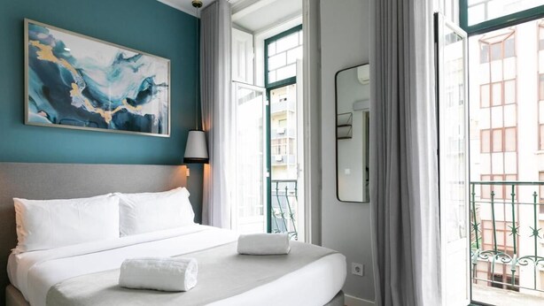 Habitación de hotel con una cama grande, un cuadro en la pared, una ventana con vistas a edificios y un balcón con barandilla.