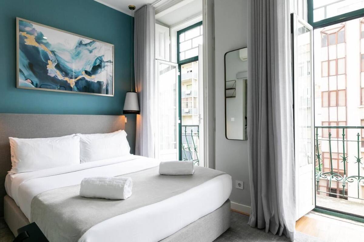 A hotel room with a large bed, a painting on the wall, a window with a view of buildings, and a balcony with a railing.