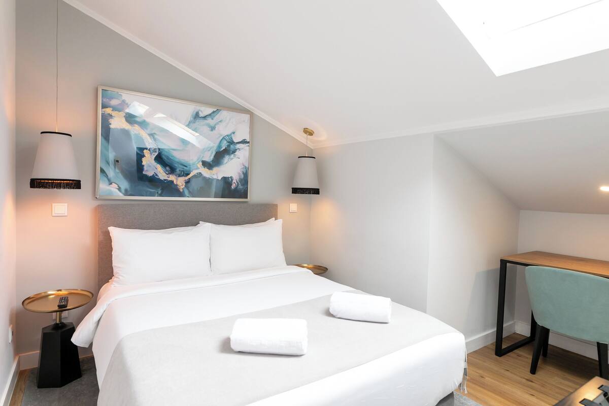 A modern hotel room with a neatly made bed, a desk, and a large abstract painting on the wall.