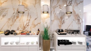 Un moderno vestíbulo de hotel con paredes de mármol, una planta central y una zona de bufé con diferentes alimentos y salsas.