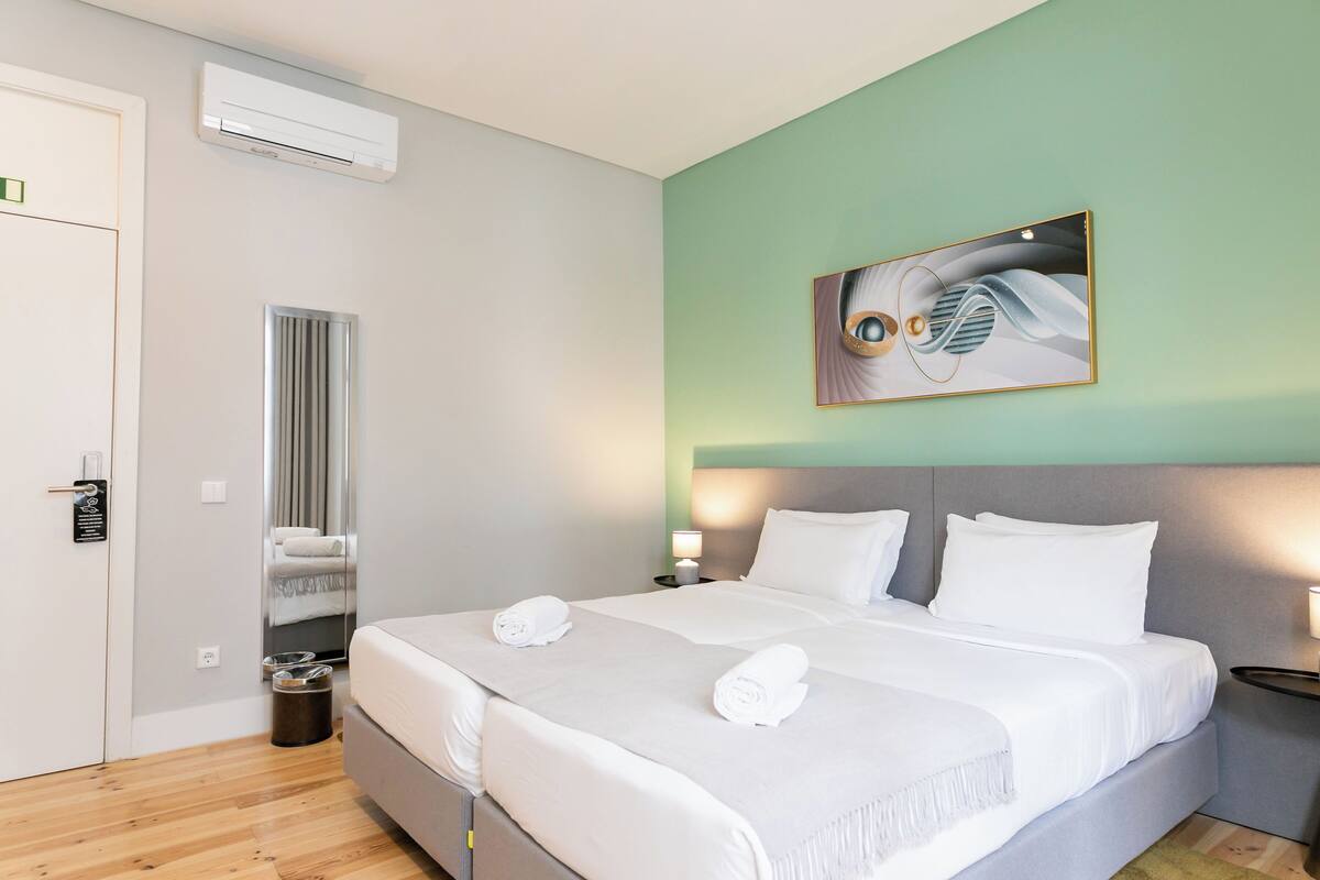 A hotel room with a bed, bedside tables, an air conditioning unit, a wall-mounted artwork, and a mirror.