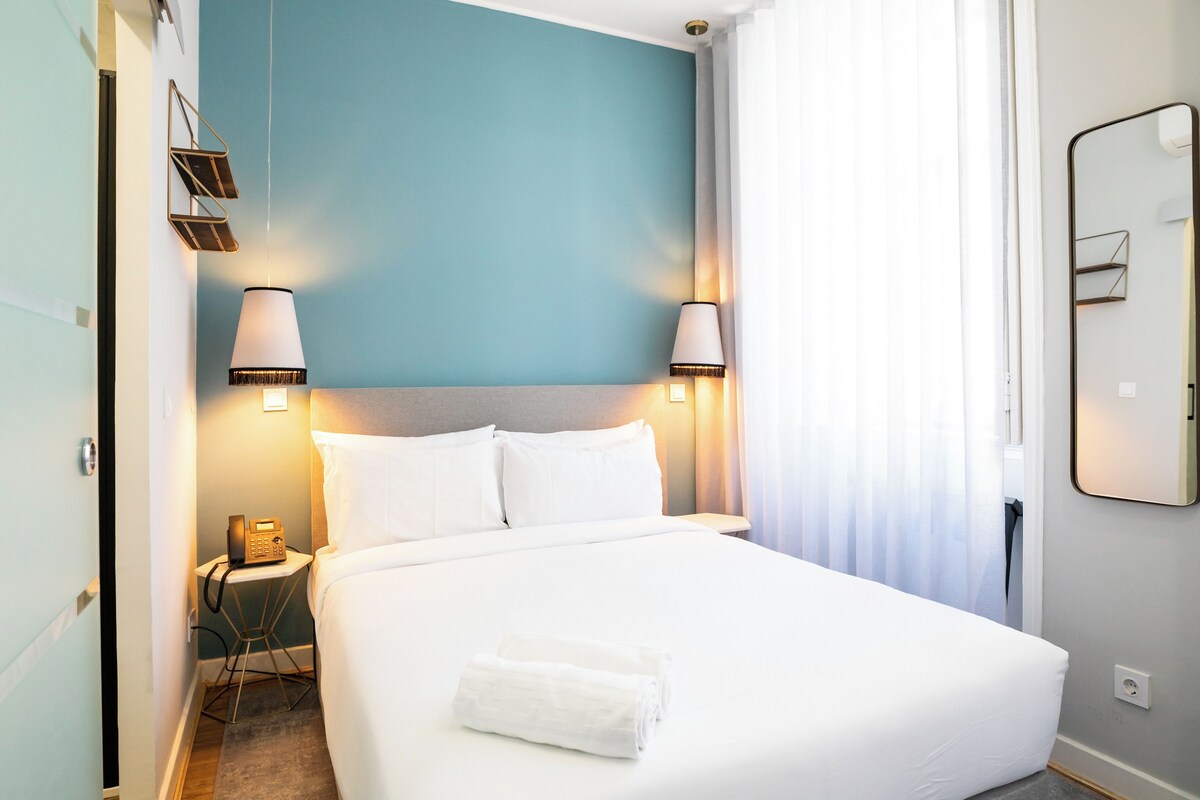 A hotel room with a bed, two bedside lamps, a telephone, and a folded towel on the bed.