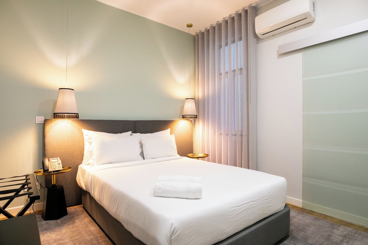 A neatly made bed with white linens, two bedside lamps, a bedside phone, and an air conditioning unit on the wall.