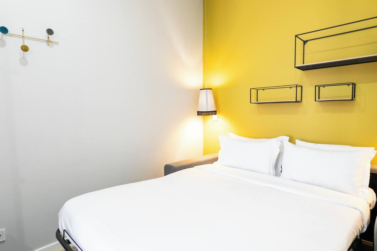 A hotel room with a bed, a nightstand, a wall-mounted lamp, and a shelf with three picture frames.