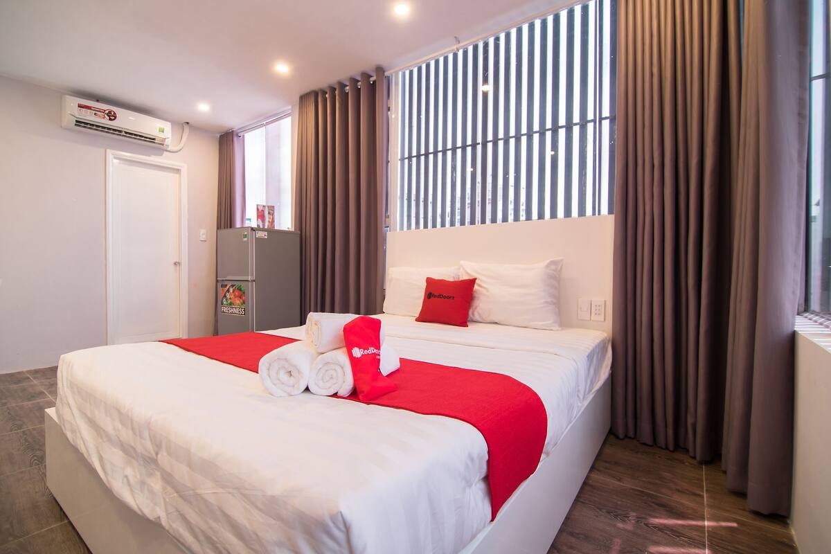 A hotel room with a large bed, a refrigerator, a window with curtains, and an air conditioning unit.