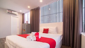 A hotel room with a large bed, a refrigerator, a window with curtains, and an air conditioning unit.